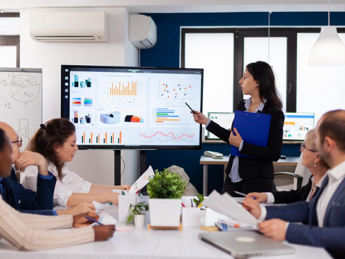 Young team leader in big corporation briefing coworkers pointing at graph meeting. Corporate staff discussing new business application with colleagues looking at screen